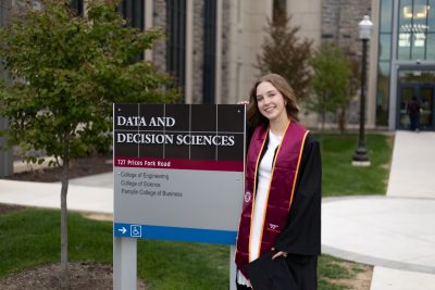 Student in graduation regalia stands next to sign for the Data and Decision Sciences building.