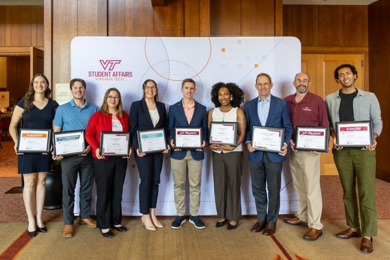 Nine smiling award recipients stand in front of a Virginia Tech Student Affairs-branded backdrop. They are all dressed in professional attire, and each holds their award certificate in front of them.