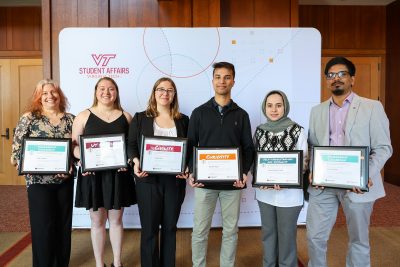Six people stand smiling and holding variously colored award certificates, before a backdrop that reads "Virginia Tech Student Affairs."